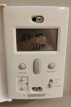 A beige thermostat on a wall, displaying the current time, the number "7.0" indicating the relative temperature, and a few manual control buttons to program the device.