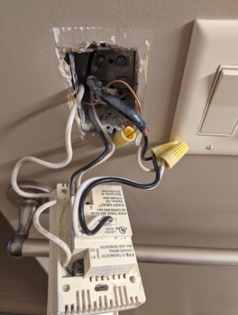 A beige thermostat pulled out of the wall; black and white wires run into it. At the top of the device, bottom of the photo, a black and white small gauge wire run in from the thermistor as a temperature sensor. In the middle, heavier wires carry the AC load into the resistive heating of the floor. And at the bottom, black and white wires provide the AC power input.